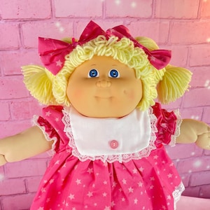 May include: A Cabbage Patch Kid doll with blonde hair, blue eyes, and a pink dress with white stars. The doll is wearing white shoes and a white bib with a pink bow.