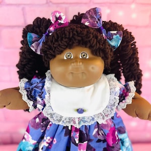 May include: A Cabbage Patch Kid doll with brown curly hair, wearing a blue floral dress with white lace trim and a white bib. The doll has brown eyes and a brown complexion.