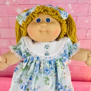 May include: A vintage doll with yellow yarn hair and blue eyes. The doll wears a light blue dress with a floral pattern of blue roses, a white collar, and lace trim. The doll's outfit includes matching lace-trimmed bloomers and a hair bow.