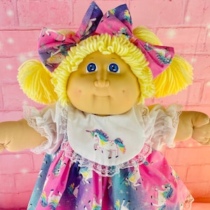 以下が含まれることがあります： 黄色い毛糸の髪、青い目、丸い顔をしたヴィンテージ人形。人形は、ユニコーンの白いよだれかけ、ユニコーン模様のピンクと青のドレス、大きなリボン、ピンクの靴を着用しています。コレクションアイテムです。
