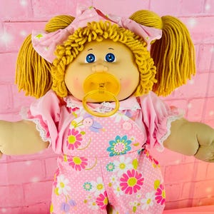 May include: A vintage doll with blonde yarn hair styled in pigtails and a pink bow. The doll wears a pink outfit with a floral pattern, a matching bonnet, and a pacifier. The doll has blue eyes and a neutral expression.