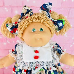 May include: A Cabbage Patch Kid doll wearing a blue and white dress with a Christmas dog print. The doll has blonde hair in pigtails with matching bows, green eyes, and red shoes.