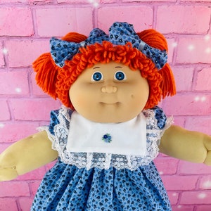 May include: A Cabbage Patch Kid doll with red curly hair, wearing a blue and white floral dress, white lace collar, and brown shoes.