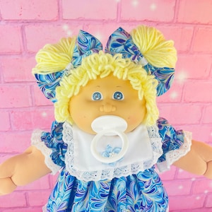 以下が含まれることがあります： 金髪で青い目をしたキャベツパッチキッズのドール。白いレースのトリムが付いた青と紫のフローラルドレスと紫色の靴を履いています。