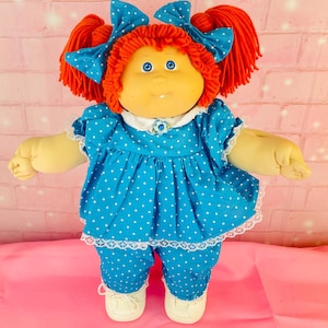 May include: A soft-sculpture doll with bright orange yarn hair styled in pigtails with blue polka-dot bows. The doll has blue eyes, a small tooth, and wears a blue polka-dot dress with a white collar and lace trim, matching pants, and white shoes.