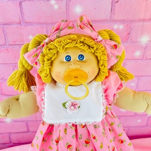 May include: A Cabbage Patch Kid doll wearing a pink dress with a white bib and a pink headband with a floral pattern. The doll has blonde curly hair and blue eyes. The doll is holding a yellow dummy.