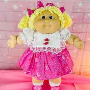 May include: A doll with yarn blonde hair styled in pigtails with pink polka dot bows. It wears a white lace-trimmed blouse with two small red rose accents and a bright pink skirt featuring a white dragonfly pattern, white socks, and pink shoes.