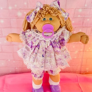 May include: A doll with tan skin, yarn hair in pigtails with matching purple floral bows, and green eyes. It wears a purple and white floral dress with lace trim, matching bloomers, white socks, and purple shoes, with a pink pacifier in its mouth.