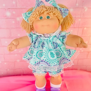 Peut inclure: Une poupée aux cheveux blonds en laine coiffés en couettes avec des nœuds à motifs bleu sarcelle. Elle porte une robe à imprimé mandala bleu sarcelle et violet avec bordure en dentelle blanche, un bloomer assorti, des chaussettes et chaussures violettes.