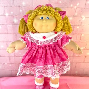 May include: A doll with blonde yarn hair styled in pigtails, blue eyes, and rosy cheeks. The doll wears a bright pink dress with a floral pattern, a white collar with lace trim and small red flower accents, white socks, and shiny pink shoes.