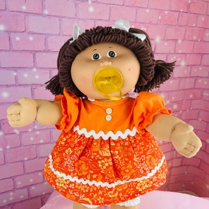 May include: A Cabbage Patch Kid doll with brown hair wearing a bright orange dress with white trim and a floral pattern. The doll is holding a yellow pacifier and is wearing orange shoes.