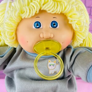 Op de afbeelding: Een Cabbage Patch Kid pop met blond garenhaar, die een grijs sweatshirt draagt met een blauwe rand en een kattenprint. De pop houdt een gele speen vast.