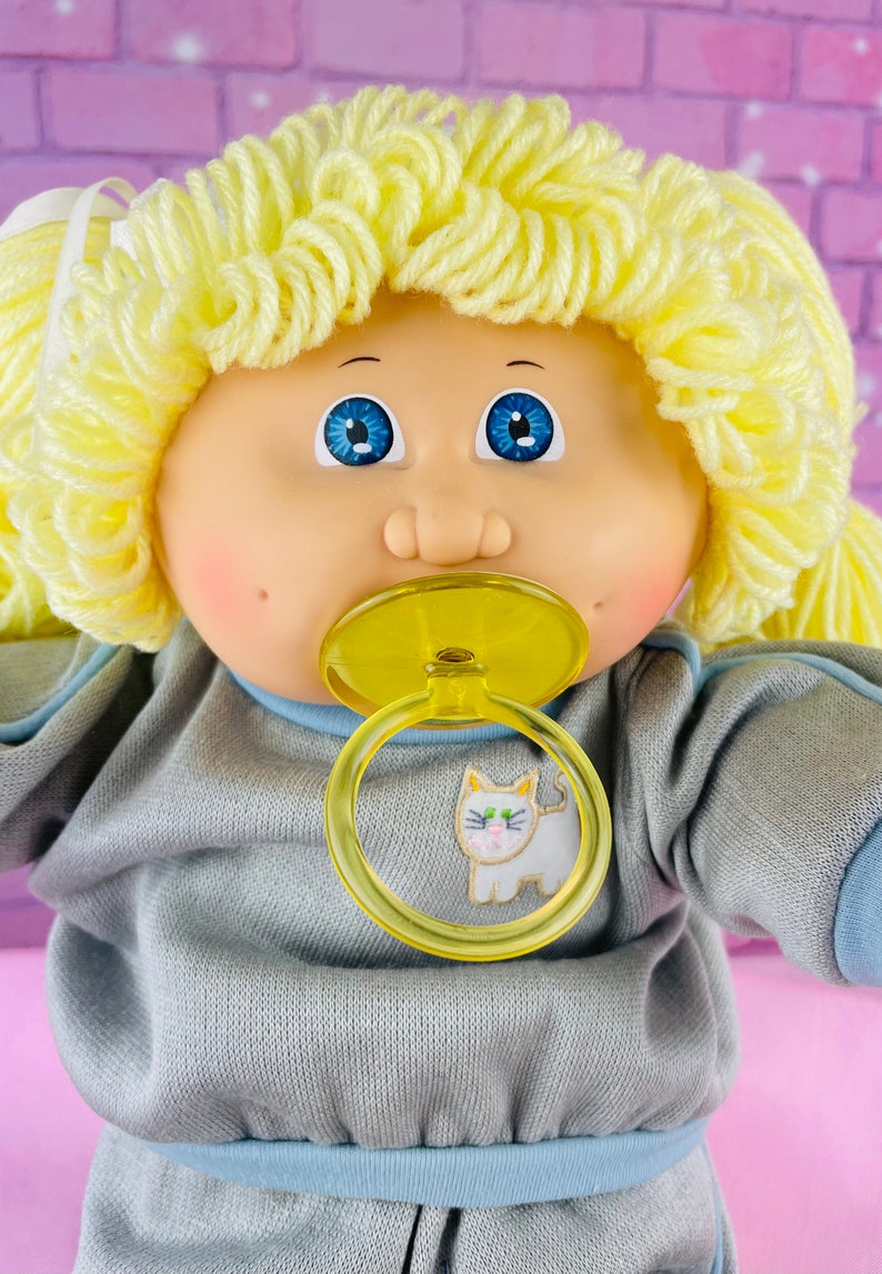 Op de afbeelding: Een Cabbage Patch Kid pop met blond garenhaar, die een grijs sweatshirt draagt met een blauwe rand en een kattenprint. De pop houdt een gele speen vast.