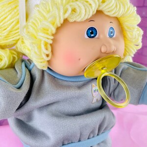 Op de afbeelding: Een Cabbage Patch Kid pop met blond garen haar, blauwe ogen en een speen. De pop draagt een grijs sweatshirt met een blauwe rand en een wit kattenontwerp.