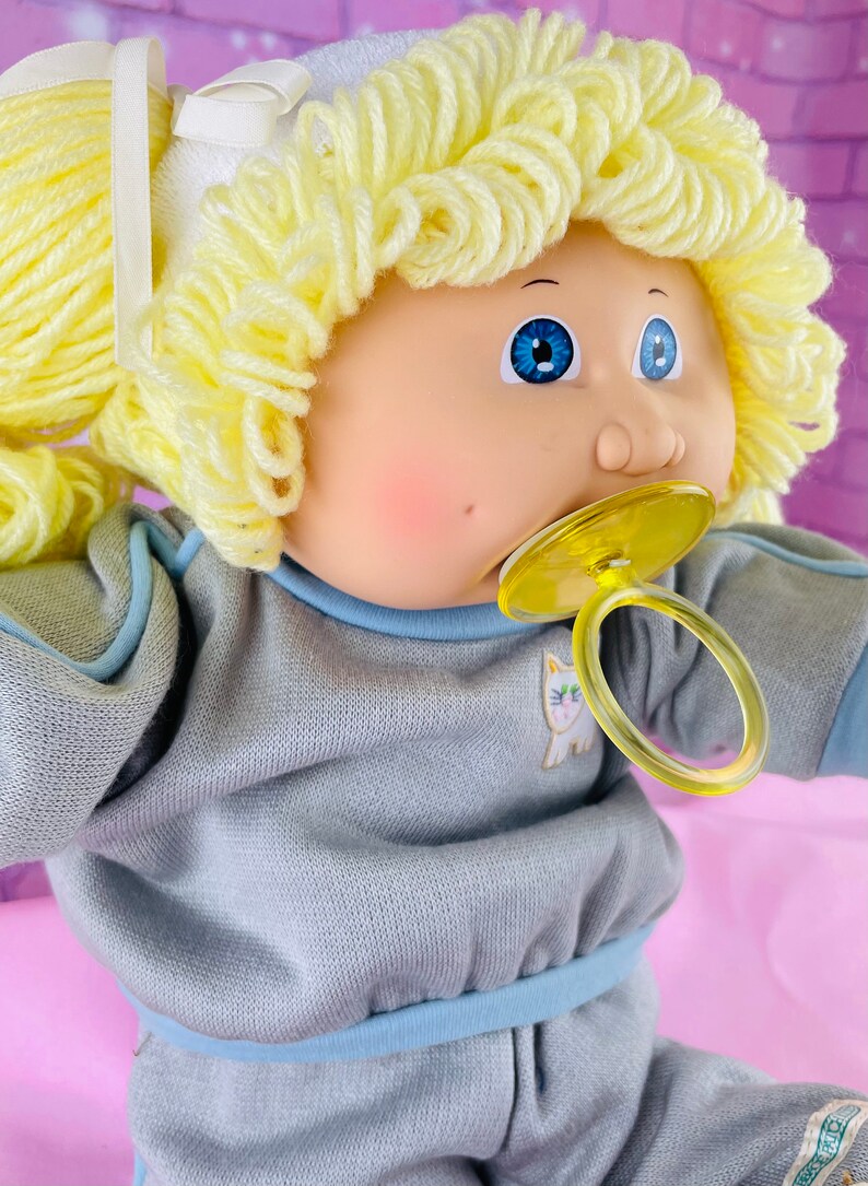 Op de afbeelding: Een Cabbage Patch Kid pop met blond garen haar, blauwe ogen en een speen. De pop draagt een grijs sweatshirt met een blauwe rand en een wit kattenontwerp.