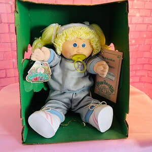 Op de afbeelding: Een Cabbage Patch Kid pop met blond haar, die een blauw-grijs trainingspak en witte sneakers met roze strepen draagt. De pop zit in een groene doos met een Cabbage Patch Kids geboortecertificaat en papieren.