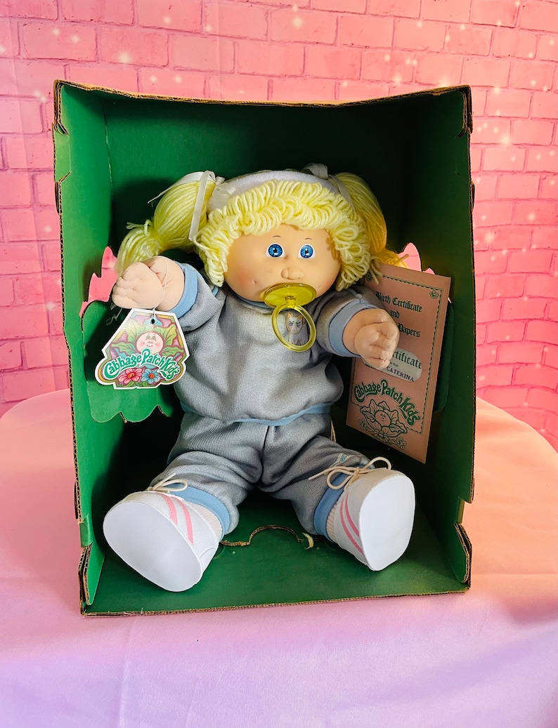 Op de afbeelding: Een Cabbage Patch Kid pop met blond haar, die een blauw-grijs trainingspak en witte sneakers met roze strepen draagt. De pop zit in een groene doos met een Cabbage Patch Kids geboortecertificaat en papieren.
