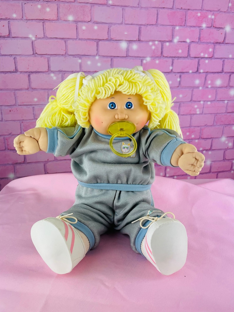 Op de afbeelding: Een Cabbage Patch Kid pop met blond garen haar, die een grijs trainingspak draagt met blauwe accenten en witte sneakers met roze strepen. De pop houdt een gele speen vast met een kattenontwerp.