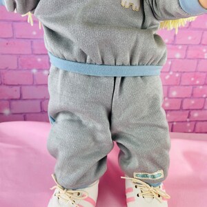 Op de afbeelding: Een grijze en blauwe babypop outfit met een kattenapplicatie en witte sneakers met roze strepen. De pop draagt ​​een grijs sweatshirt met een blauwe rand en een bijpassende broek. De schoenen van de pop hebben de tekst "CAROLE B. KIDS" aan de zijkant.