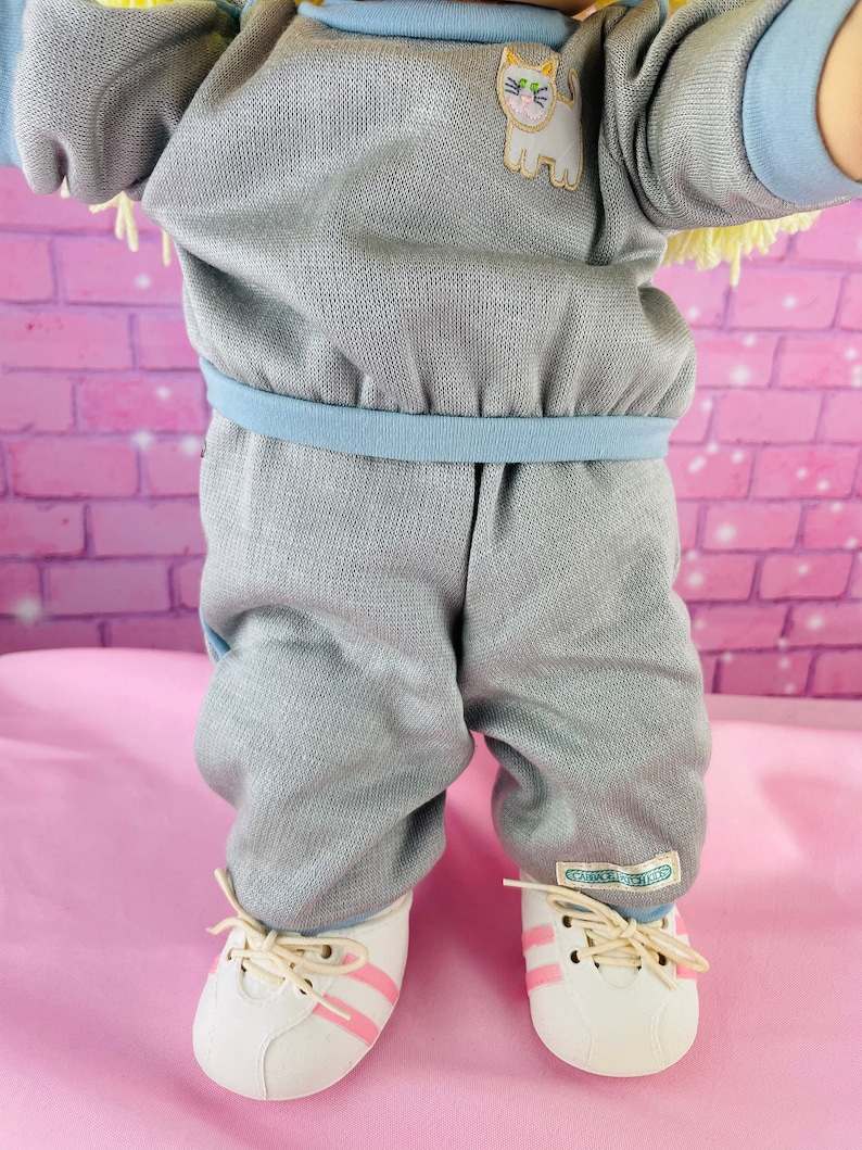 Op de afbeelding: Een grijze en blauwe babypop outfit met een kattenapplicatie en witte sneakers met roze strepen. De pop draagt ​​een grijs sweatshirt met een blauwe rand en een bijpassende broek. De schoenen van de pop hebben de tekst "CAROLE B. KIDS" aan de zijkant.