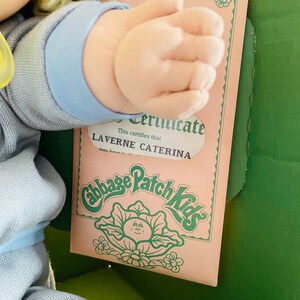 Op de afbeelding: Een Cabbage Patch Kids pop met een blauwe outfit en een certificaat met de tekst: "Dit certificaat verklaart dat Laverne Caterna geboren is op..."