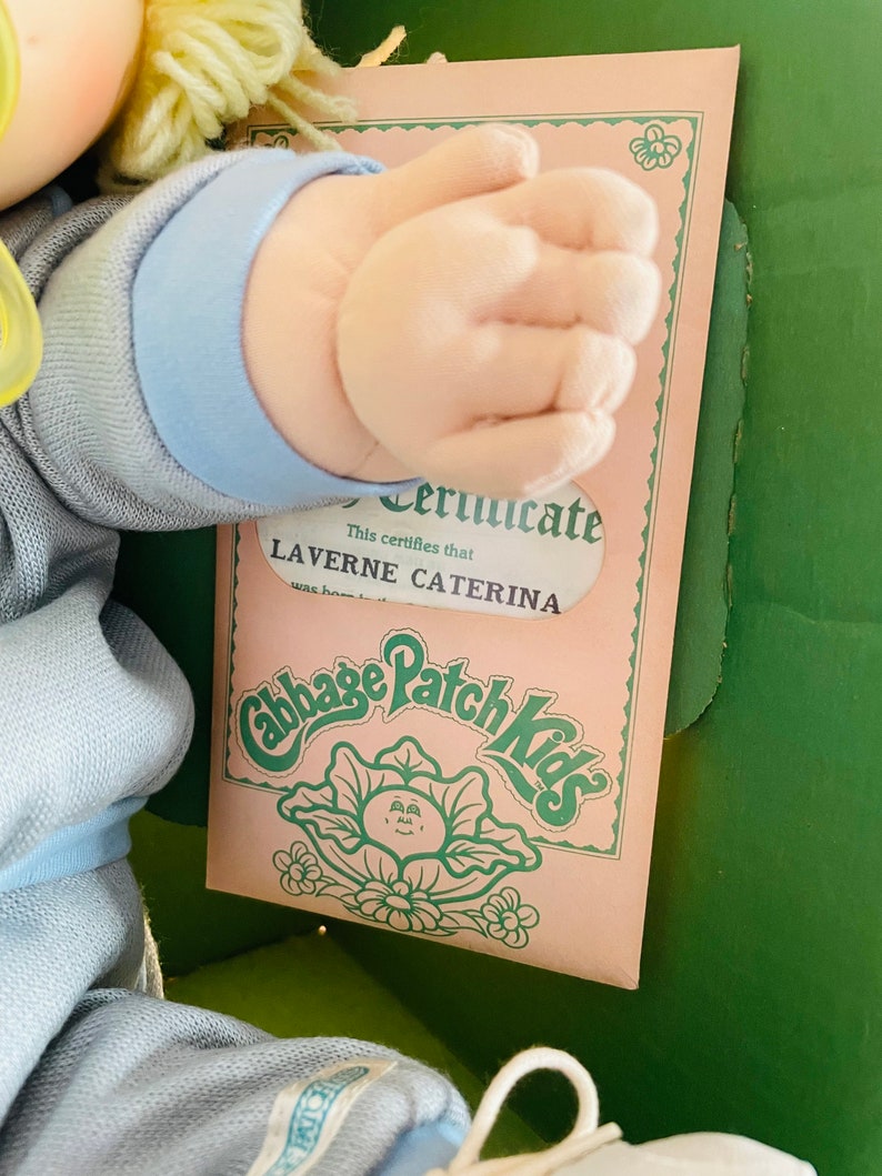 Op de afbeelding: Een Cabbage Patch Kids pop met een blauwe outfit en een certificaat met de tekst: "Dit certificaat verklaart dat Laverne Caterna geboren is op..."