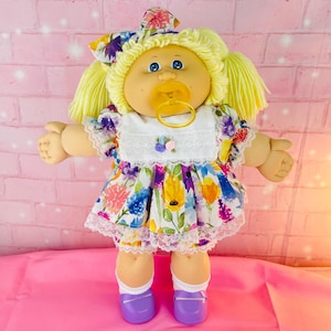 May include: A doll with yellow yarn hair in pigtails, wearing a floral print dress with a white lace-trimmed bib and a matching hair bow. The doll has blue eyes, a yellow pacifier, white socks, and purple shoes, featuring a soft fabric body.