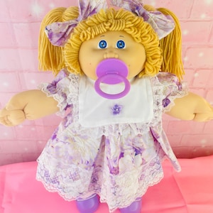 Peut inclure: Une poupée aux cheveux en laine blonde coiffés en couettes avec des nœuds à motifs floraux violets assortis. La poupée a des yeux bleus, une tétine violette et porte un bavoir blanc sur une robe à fleurs violettes avec de la dentelle blanche.