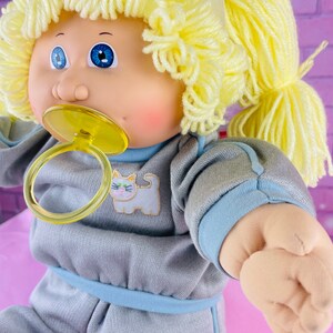 Op de afbeelding: Een Cabbage Patch Kid pop met blond garen haar in paardenstaarten, die een grijze en blauwe slaapzak draagt met een kattenontwerp. De pop heeft blauwe ogen en houdt een gele speen vast.