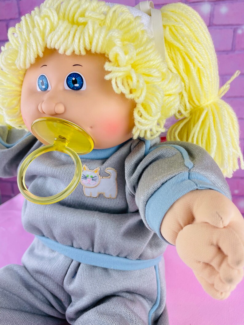 Op de afbeelding: Een Cabbage Patch Kid pop met blond garen haar in paardenstaarten, die een grijze en blauwe slaapzak draagt met een kattenontwerp. De pop heeft blauwe ogen en houdt een gele speen vast.