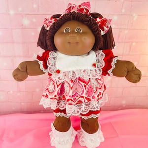 May include: A doll with dark brown yarn hair, brown eyes, and a dimpled face. The doll wears a red dress with a white heart-patterned skirt, white lace trim, a white bib with small red roses, matching hair bows, and white lace-trimmed socks.