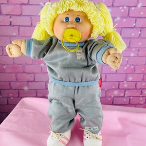 Op de afbeelding: Een Cabbage Patch Kid pop met blond haar, blauwe ogen en een speen. De pop draagt een grijs trainingspak met blauwe accenten en witte sneakers met roze strepen.