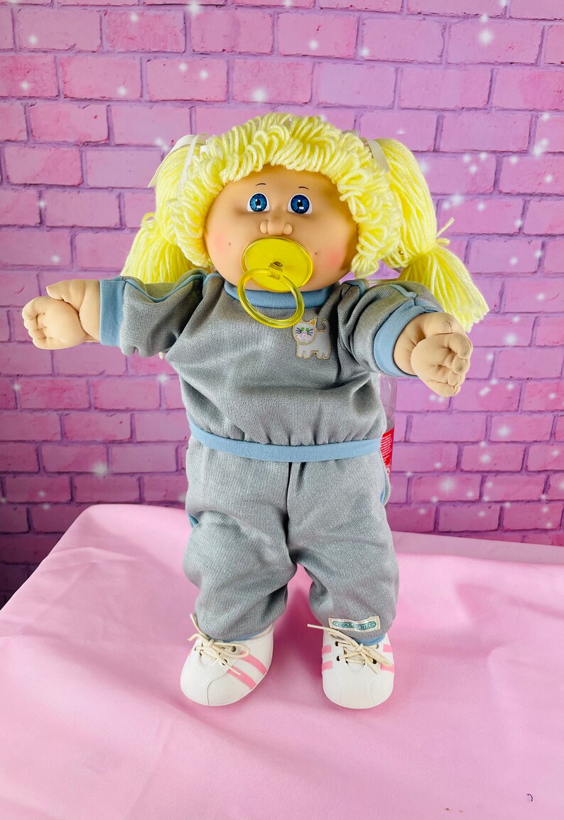 Op de afbeelding: Een Cabbage Patch Kid pop met blond haar, blauwe ogen en een speen. De pop draagt een grijs trainingspak met blauwe accenten en witte sneakers met roze strepen.