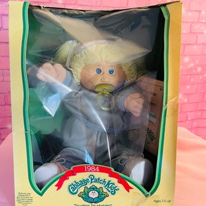 Op de afbeelding: Een vintage Cabbage Patch Kids pop in een blauwe outfit met een speen, zittend in een doos met de tekst "1984 Cabbage Patch Kids" en "Ik ben uniek. Ik ben adopteerbaar!" De doos is groen en geel met een witte achtergrond.