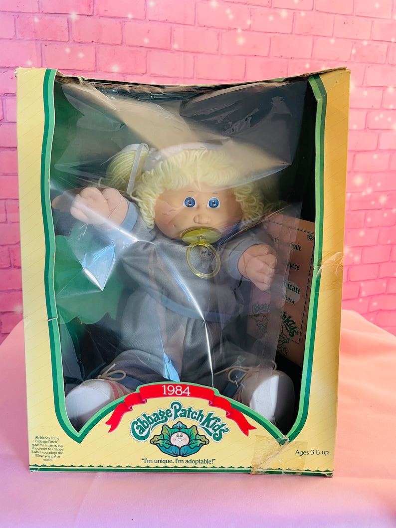 Op de afbeelding: Een vintage Cabbage Patch Kids pop in een blauwe outfit met een speen, zittend in een doos met de tekst "1984 Cabbage Patch Kids" en "Ik ben uniek. Ik ben adopteerbaar!" De doos is groen en geel met een witte achtergrond.