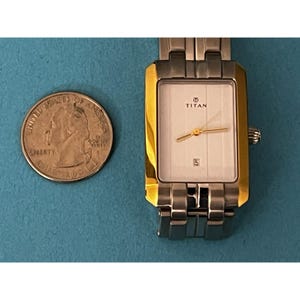May include: A rectangular Titan wristwatch with a silver band and gold-toned bezel. The watch face is white with gold hands and the date displayed at the 6 o'clock position. A U.S. quarter is next to the watch.