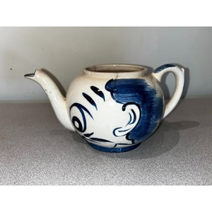 May include: A white ceramic teapot with a blue and white cartoon face painted on the side. The teapot has a spout and a handle.