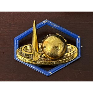 May include: A gold-colored pin with a blue hexagonal frame. The pin depicts the 1964 New York World's Fair logo, featuring a stylized representation of the Unisphere and the New York State Pavilion.
