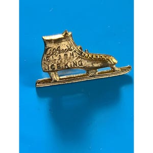 May include: A gold-colored ice skate pin with the text "Alfred's Flash Ice King" on the side. The pin is detailed with a blade and laces, set against a bright blue background.