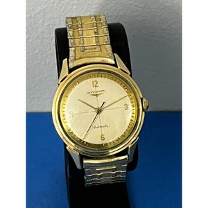 May include: A gold-toned Longines automatic wristwatch with a cream-colored face. The watch features gold hands and hour markers, with the number 12 at the top. The watch band is a combination of gold and silver-toned metal. The word "Longines" is printed on the watch face.