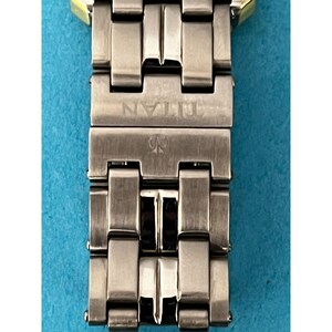 May include: Close-up of a silver-toned watch band with the brand name "TITAN" engraved on the clasp. The band is made of rectangular metal links with a gold-colored accent at the top. The watch band is on a blue background.