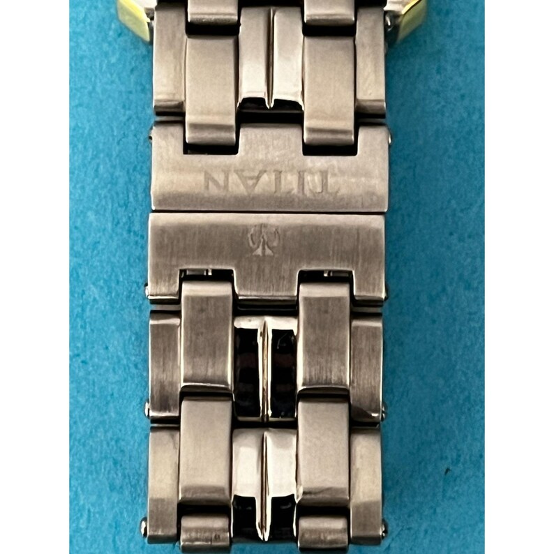 May include: Close-up of a silver-toned watch band with the brand name "TITAN" engraved on the clasp. The band is made of rectangular metal links with a gold-colored accent at the top. The watch band is on a blue background.