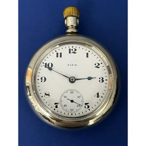 May include: A silver Elgin pocket watch with a white face and black numerals. The watch has a small seconds dial and a gold-colored crown. The hands are black, and the watch is set against a blue background.