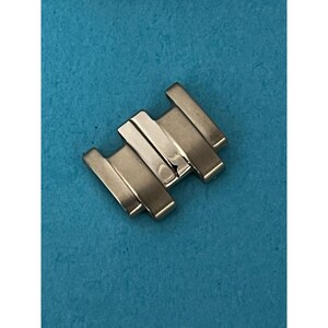 May include: A silver-toned watch clasp, featuring a series of rectangular links. The clasp is designed with a folding mechanism, and is set against a solid blue background. The metal has a polished finish.
