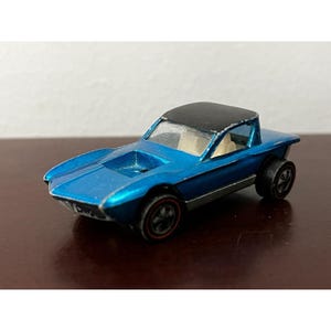 May include: A blue toy car with a black roof and a white interior. The car has a long, low profile and a distinctive design. It is a vintage Hot Wheels car.