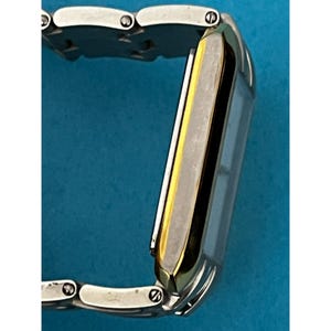 May include: A rectangular wristwatch with a silver band and a gold-colored bezel. The watch face is a light color, and the band is made of multiple small, connected links. The watch is shown against a blue background.