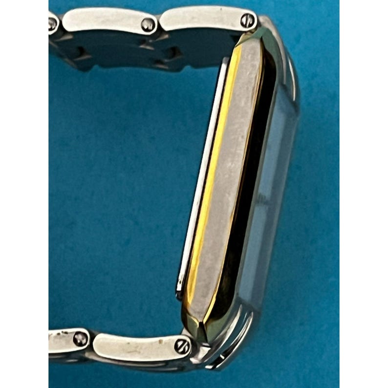May include: A rectangular wristwatch with a silver band and a gold-colored bezel. The watch face is a light color, and the band is made of multiple small, connected links. The watch is shown against a blue background.