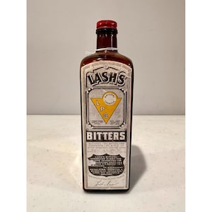 May include: A vintage brown glass bottle of Lash's Bitters with a red cap. The label features the text "LASH'S" and "BITTERS" in a decorative font, along with other text. The bottle is rectangular.