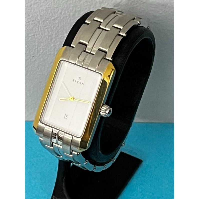May include: A rectangular Titan watch with a silver band and a gold-colored bezel. The watch face is white with gold hands and the number 15 at the bottom. The watch is displayed on a black stand.