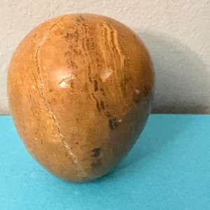 May include: A polished, egg-shaped stone with a warm, brown color and natural grain patterns. The stone has a smooth, glossy surface and a crack running down its side. The object is set against a blue surface.